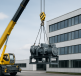 A crane lifting heavy machinery in an industrial setting with a modern building in the background.