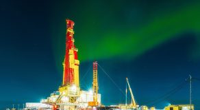 A brightly lit oil drilling rig under an aurora-filled night sky.