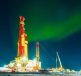 A brightly lit oil drilling rig under an aurora-filled night sky.